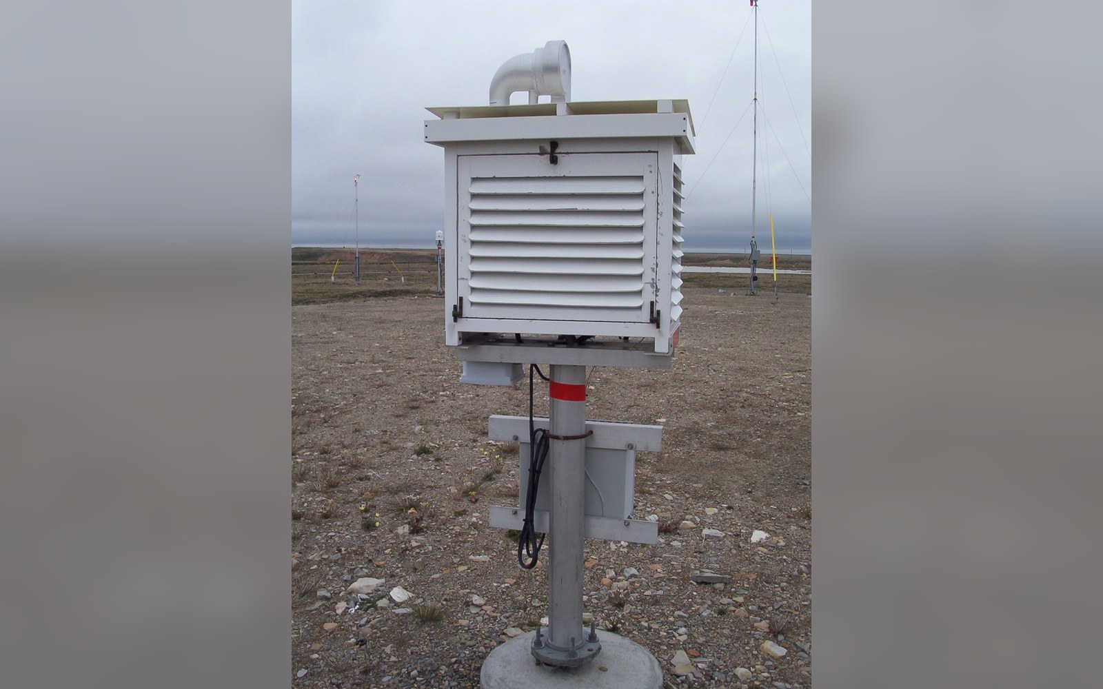 A louvered weather instrument box mounted on a pole in a gravel field